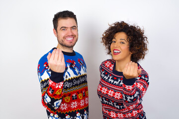 Young couple wearing Christmas sweater standing against white wall inviting to come with hand. Happy that you came