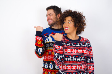 Charming Young couple wearing Christmas sweater standing against white wall looking at copy space having advertisements