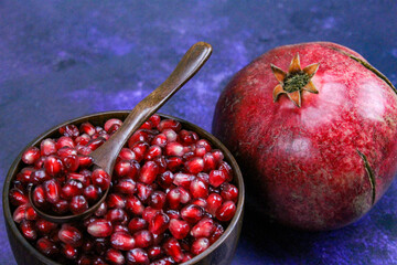 A bowl full of pomegranate seeds with a wooden spoon on it. A whole ripe pomegranate next to the bowl. Seeds and pomegranate are on a blue background.