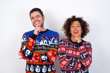 Optimistic Young couple wearing Christmas sweater standing against white wall keeps hands partly crossed and hand under chin, looks at camera with pleasure. Happy emotions concept.