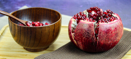 Wooden bowl containing wooden spoon and red pomegranate seeds. Pomegranate fruit open divided into five parts held together on a napkin / cloth on wooden tray on a blue background. Fruit pomegranate.