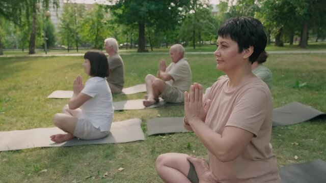 Panning of middle-aged attractive woman with short dark hair sitting on mat on grass in park and doing yoga with several senior people on background