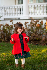 toddler girl in dark green dress and red coat standing outside in a patch of sun