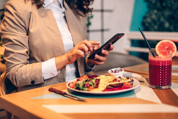 A middle-aged woman is sitting in a restaurant, typing a message on her mobile phone and enjoys a healthy meal and fresh beetroot and grapefruit juice.