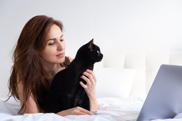 attractive woman using laptop with her black cat, laying together in white bedroom. life with pet.