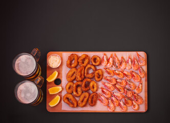 Beer, fried squid rings and shrimps on cutting board on dark background. Top view, copy space.