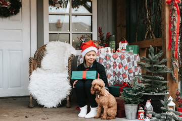 a boy in Santa hats with a dog sit on the porch of a house decorated for Christmas and boxes with gifts in his hands, the concept of a family holiday of Christmas and New year