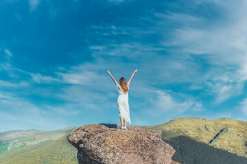 Woman from behind with white dress and her arms raised on top of a large rock on a valley landscape...