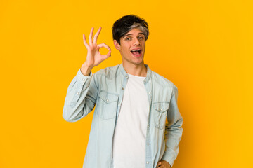 Young skinny hispanic man winks an eye and holds an okay gesture with hand.