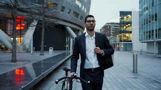 Mixed race businessman walking bicycle wearing backpack leaving work after long day 