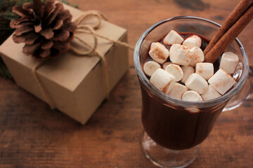 Christmas hot chocolate with marshmallows and cinnamon in a glass cup and craft gift box on a wooden table. Hot winter drink on wood background.
