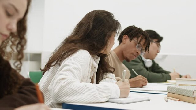 Chest up side view of Mixed-Race girl peeping in notebook of African desk mate. Diverse students sitting by desks in bright classroom, taking notes on lecture