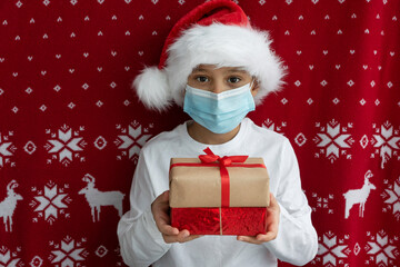 white caucasian boy in santas hat and face medical mask holds two gift boxes tired with ribbons on red christmas background. Holiday concept