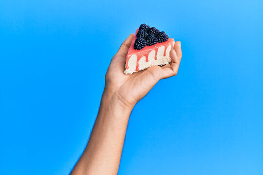 Hand of hispanic man holding slice of cheesecake over isolated bue background.