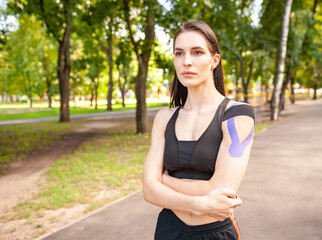 Portrait of attractive muscular brunette woman wearing black sports outfit, looking at camera. Young female athlete posing with hands on waist, colorful kinesiotaping on body.