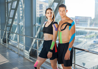 Fototapeta na wymiar Portrait of muscular brunette woman with arms crossed and man looking at camera with strong face. Young couple athletes posing indoors, colorful kinesiotaping on body, futuristic interior.