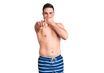 Young handsome man wearing swimwear laughing at you, pointing finger to the camera with hand over mouth, shame expression