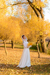 Fototapeta premium Cute girl in long white wedding dress in rural path among autumnal trees in golden hour atmosphere.