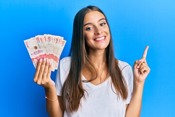 Young hispanic woman holding colombian pesos smiling happy pointing with hand and finger to the side
