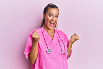 Young hispanic woman wearing doctor uniform and stethoscope celebrating surprised and amazed for success with arms raised and open eyes. winner concept.
