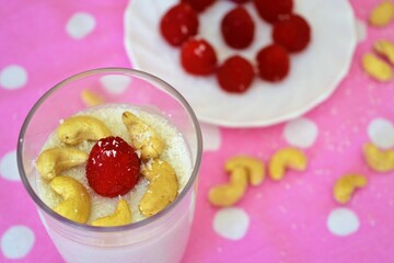 Close-up of coconut milk jelly in a glass decorated with cashews and raspberries and a plate of raspberries behind on a pink cloth with white polka dots