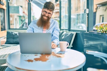 Young redhead businessman smiling happy working using laptop sitting at coffee shop terrace.