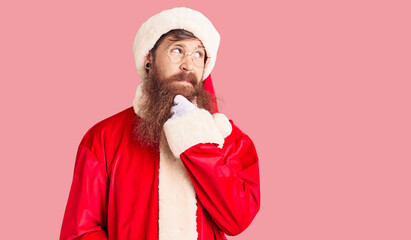 Handsome young red head man with long beard wearing santa claus costume with hand on chin thinking about question, pensive expression. smiling with thoughtful face. doubt concept.