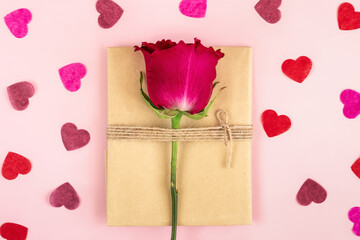Vintage gift packing in craft paper with twine and one solo pink rose and paper confetti in shape of heart against pink background.