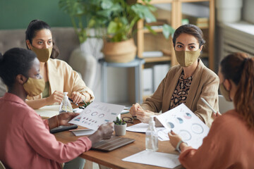 Fototapeta premium High angle view at multi-ethnic group of young businesswomen wearing masks during meeting in office, copy space