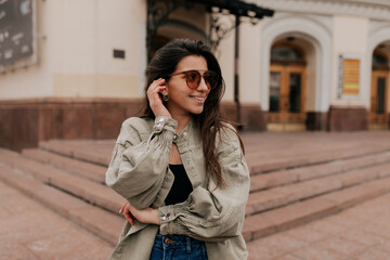 Fototapeta premium Close up portrait of a cheerful woman with glasses touching her hair on a blurred background. Photo of a fashionable girl with beautiful brown hair smiling at the camera.