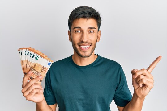 Young handsome man holding bunch of 50 euro banknotes smiling happy pointing with hand and finger to the side