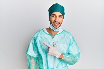 Young handsome man wearing surgeon uniform and medical mask smiling cheerful pointing with hand and finger up to the side