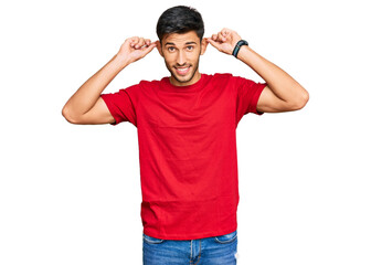 Young handsome man wearing casual red tshirt smiling pulling ears with fingers, funny gesture. audition problem