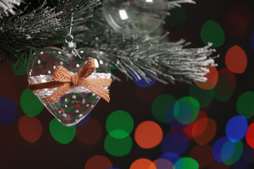 Obraz premium Heart shaped holiday bauble hanging on Christmas tree against blurred festive lights, closeup. Space for text