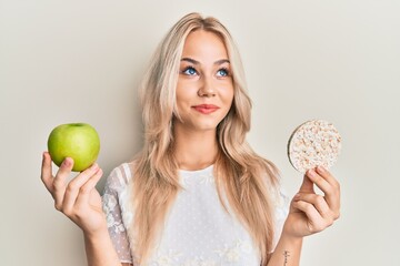 Beautiful caucasian blonde girl holding nachos and healthy green apple smiling looking to the side...