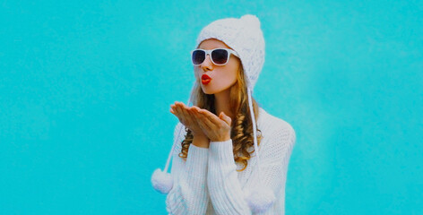 Portrait of young woman blowing red lips sending sweet air kiss wearing a white sweater and hat over blue background