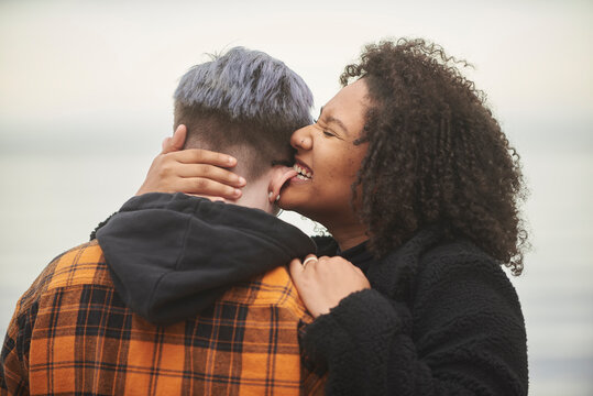 Young interracial female same sex couple embracing by water, one woman is playfully biting the other's ear.