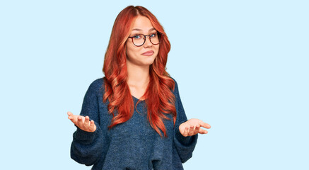 Young redhead woman wearing casual sweater and glasses clueless and confused expression with arms...