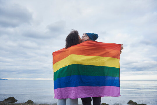 Young interracial female same sex couple kissing under a pride flag.
