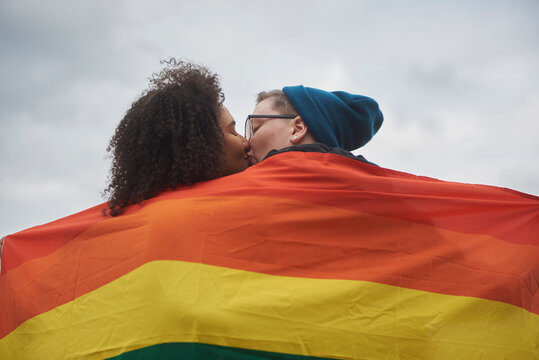 Young interracial female same sex couple kissing under a pride flag.