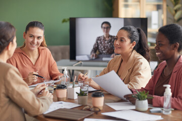 Multi-ethnic group of women discussing business project during meeting in office with remote participant in background, copy space