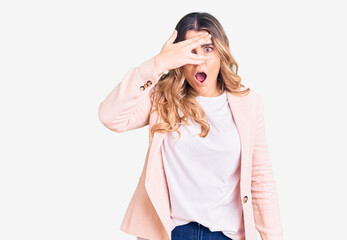 Fototapeta premium Young caucasian woman wearing business clothes peeking in shock covering face and eyes with hand, looking through fingers with embarrassed expression.