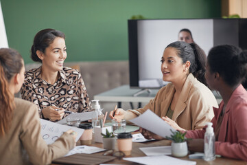 Multi-ethnic group of businesswomen discussing project during meeting in office with remote participant in background, copy space