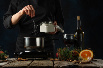 The chef pours sugar into pan for preparing mulled wine on rustic wooden table with festive composition background. Backstage of cooking hot drink with fragrant spices.