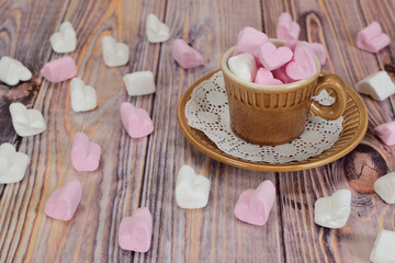 Coffee mug filled with marshmallows in the form of white and pink hearts. Remnants of marshmallows are scattered around the mug on a wooden surface.