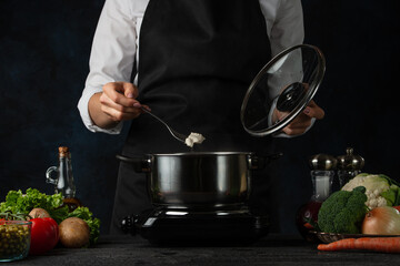 The professional chef in black apron testing hot soup on dark blue background. Backstage of preparing meal. Healthy dish. Food concept. Frozen motion. Cookbook.