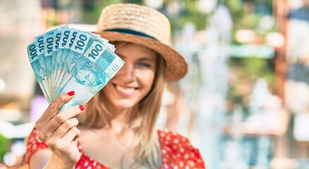 Young blonde tourist woman wearing summer style holding brazilian 100 real banknotes at the city.