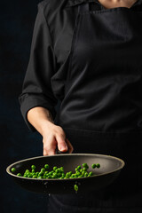 Macro shot view of chef throws up frying green peas above the pan on dark blue background. Backstage of cooking meal. Frozen motion. Food banner concept. Cookbook illustration