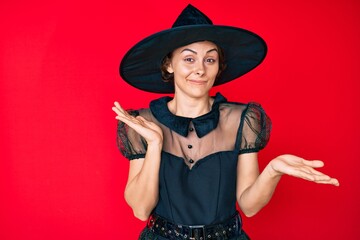 Young hispanic woman wearing witch halloween costume clueless and confused expression with arms and...