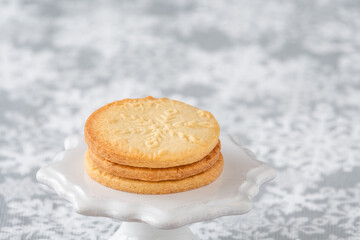 Fresh baked snowflake stamped sugar cookies on a white ceramic cake stand, and white and gray snowflake print tablecloth
